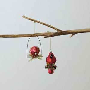Two Creative Co-op HAND PAINTED CARDINAL ORNAMENT WITH BEADS AND GLITTER pieces hang from a branch, one on a perch and one suspended vertically, both decorated with beads and berries.