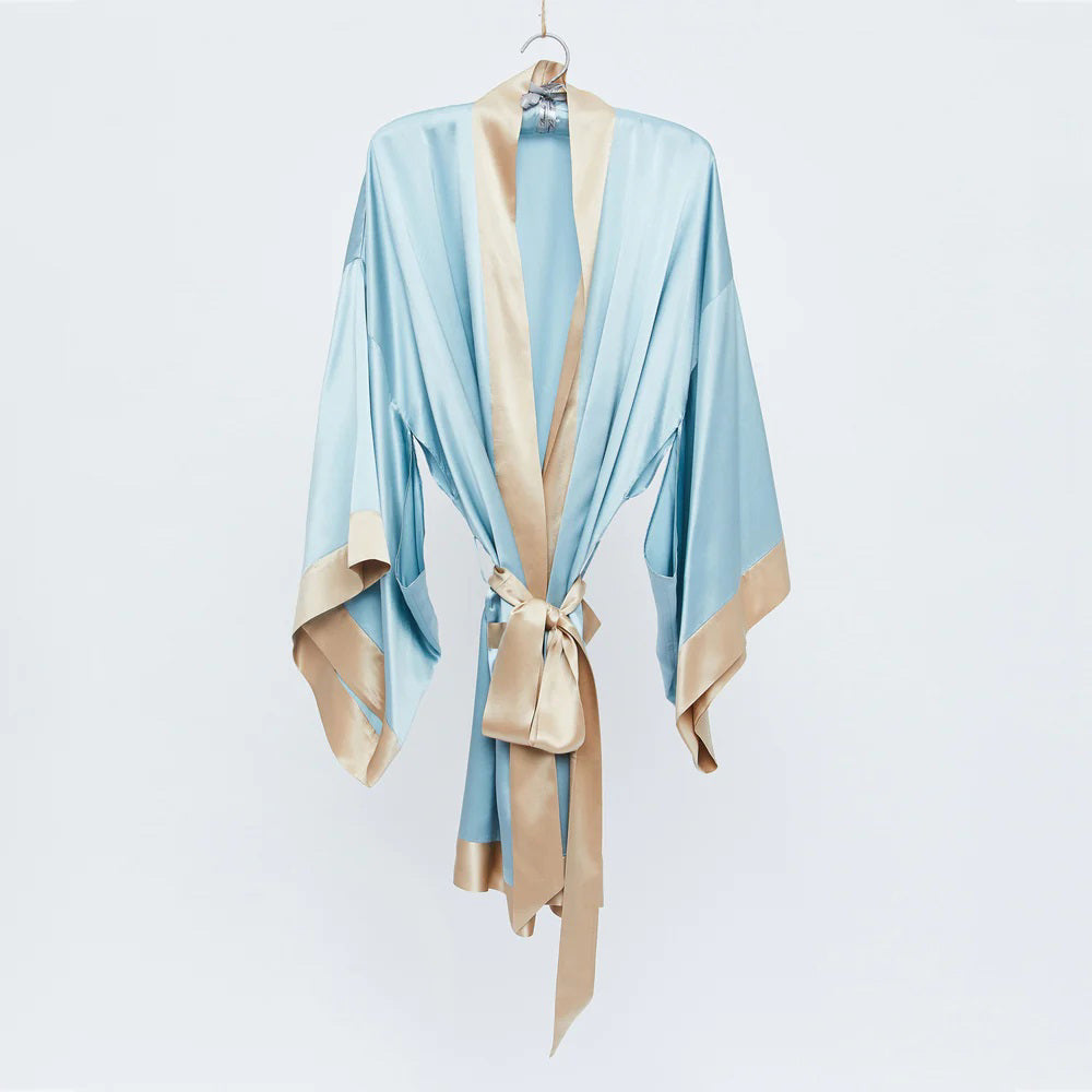 The BRANCHE - GEISHA ROBE BLUE/TOFFEE, featuring a light blue satin fabric with beige trim and a matching waist belt, is hanging on a silk hanger against a plain white background. The robe boasts wide sleeves and mitered corners, elegantly showcasing its luxurious and smooth texture.