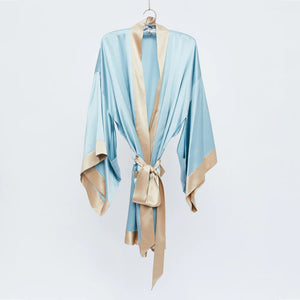 The BRANCHE - GEISHA ROBE BLUE/TOFFEE, featuring a light blue satin fabric with beige trim and a matching waist belt, is hanging on a silk hanger against a plain white background. The robe boasts wide sleeves and mitered corners, elegantly showcasing its luxurious and smooth texture.