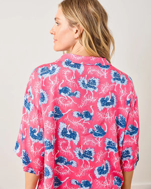 Against a neutral wall, a person stands barefoot in PRINTFRESH’s Satin Ostrich Dance Wildest Dream Pajama Set. The bright red ensemble with blue and white floral patterns features a short-sleeved button-up top and wide-legged pants, exuding comfort and style as they smile warmly.