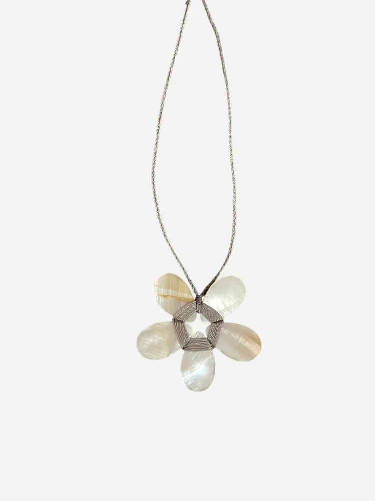 A cream-colored mannequin showcases a textured beige jacket paired with the ANN LIGHTFOOT - FLAT MOTHER OF PEARL FLOWER NECKLACE, highlighting a striking white-petaled flower pendant with a metallic center suspended from an adjustable braided cord. The background is softly blurred.