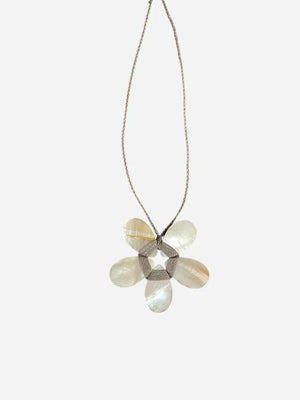 A cream-colored mannequin showcases a textured beige jacket paired with the ANN LIGHTFOOT - FLAT MOTHER OF PEARL FLOWER NECKLACE, highlighting a striking white-petaled flower pendant with a metallic center suspended from an adjustable braided cord. The background is softly blurred.