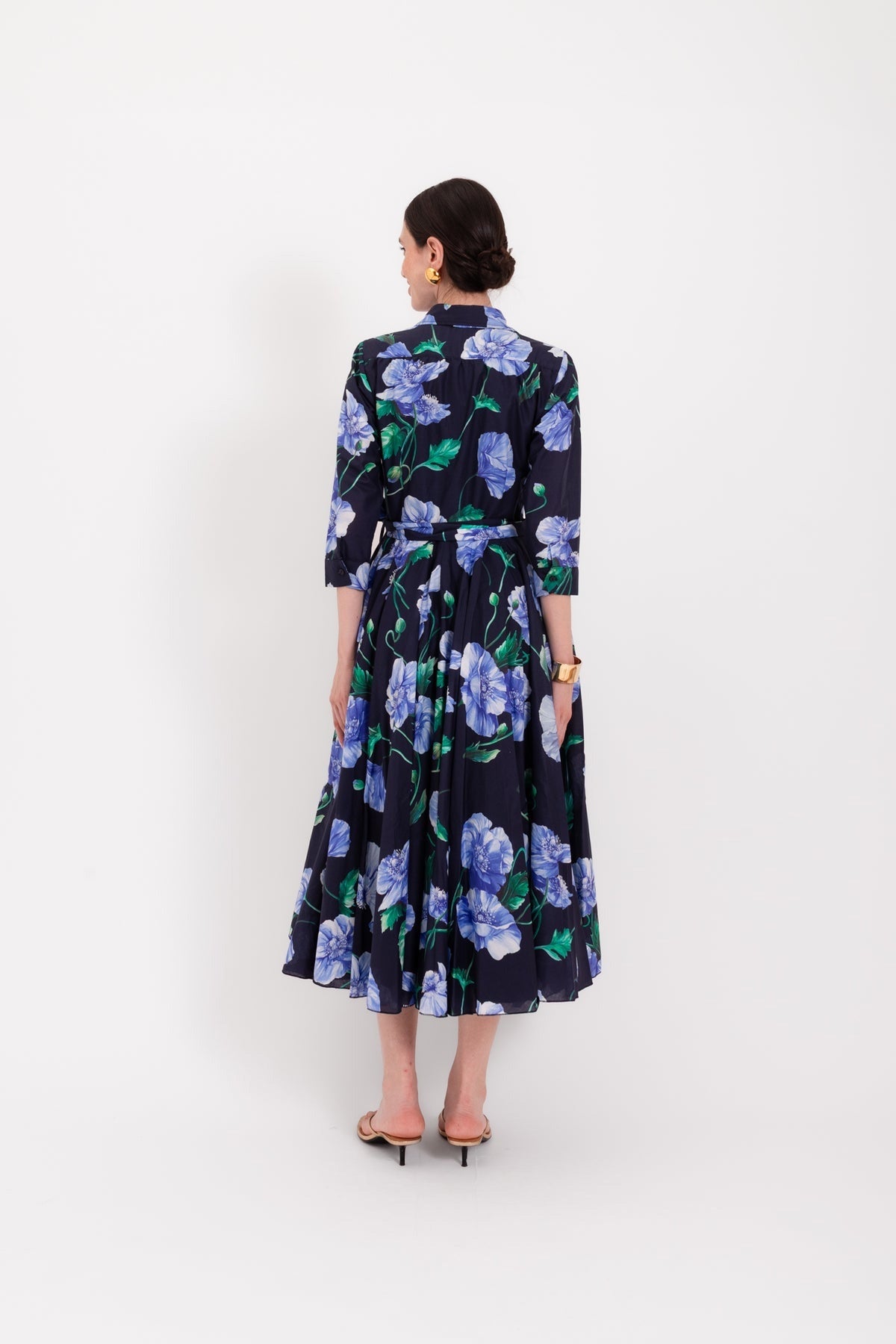 A woman stands against a white background wearing the LAVI - SPRITZ BLUE POPPIES DRESS, a navy blue, long-sleeved midi dress by LAVI featuring blue and green poppy floral prints, a belted waist, and sandals. This elegant piece is made in Italy.