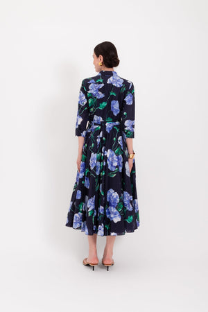 A woman stands against a white background wearing the LAVI - SPRITZ BLUE POPPIES DRESS, a navy blue, long-sleeved midi dress by LAVI featuring blue and green poppy floral prints, a belted waist, and sandals. This elegant piece is made in Italy.