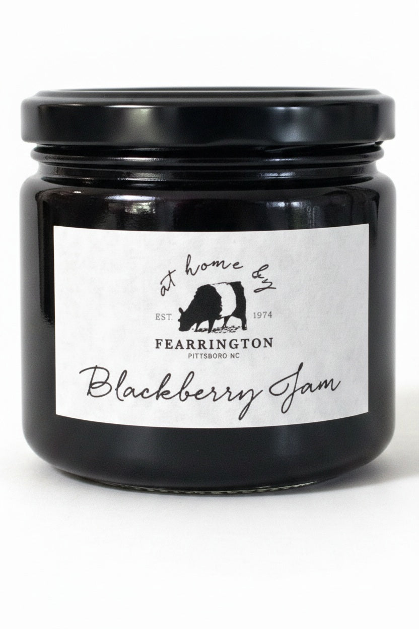The FEARRINGTON SIGNATURE COLLECTION - FEARRINGTON HOUSEMADE BLACKBERRY JAM features a stylish black and white striped ribbon tied around the lid. The label reveals ingredients including blackberry, vanilla, lemon, pectin, and sugar. This handcrafted jam is made with meticulous care and is an ideal hostess gift. Please be aware that it may contain allergens.