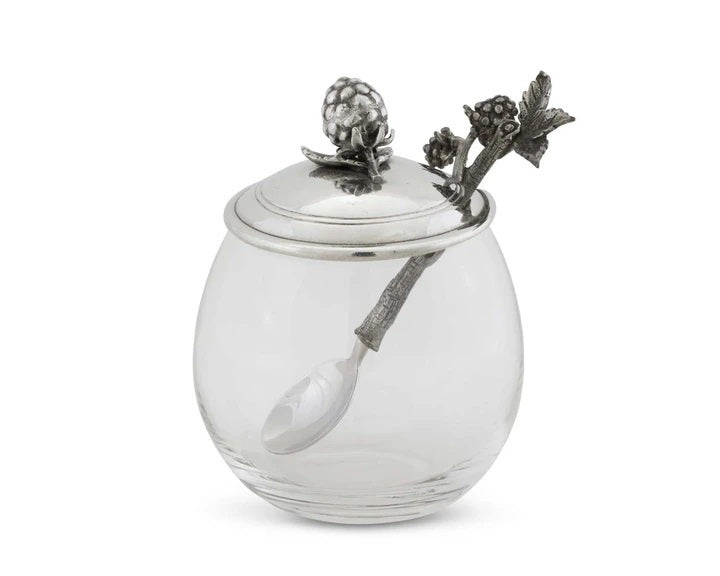 A VAGABOND HOUSE Blackberry Jam Jar, featuring a round glass design with a silver lid and decorative raspberry and leaf patterns, includes a handcrafted spoon with a branch-like handle. It&#39;s empty against a plain white backdrop.