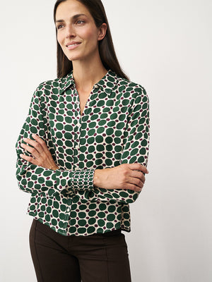 A person with straight brown hair adjusts the collar of the 0039 ITALY SIA BLOUSE, which features a geometric circle print with large green spots and small burgundy dots on cream. The side view highlights the blouse's viscose sleeve.