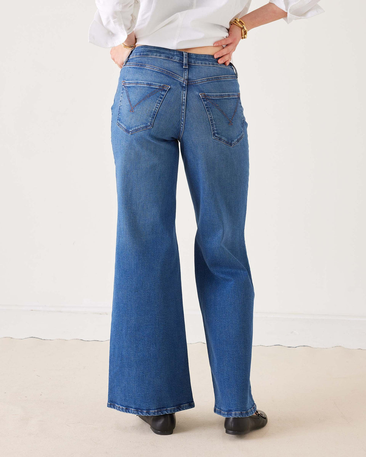 A person wearing the MERSEA Kate Wide Leg Jean in Baltic Wash, featuring four-way-stretch technology, paired with a white long-sleeve shirt with rolled cuffs, black flat shoes, and gold bracelets, stands on a light-colored floor against a plain white background.
