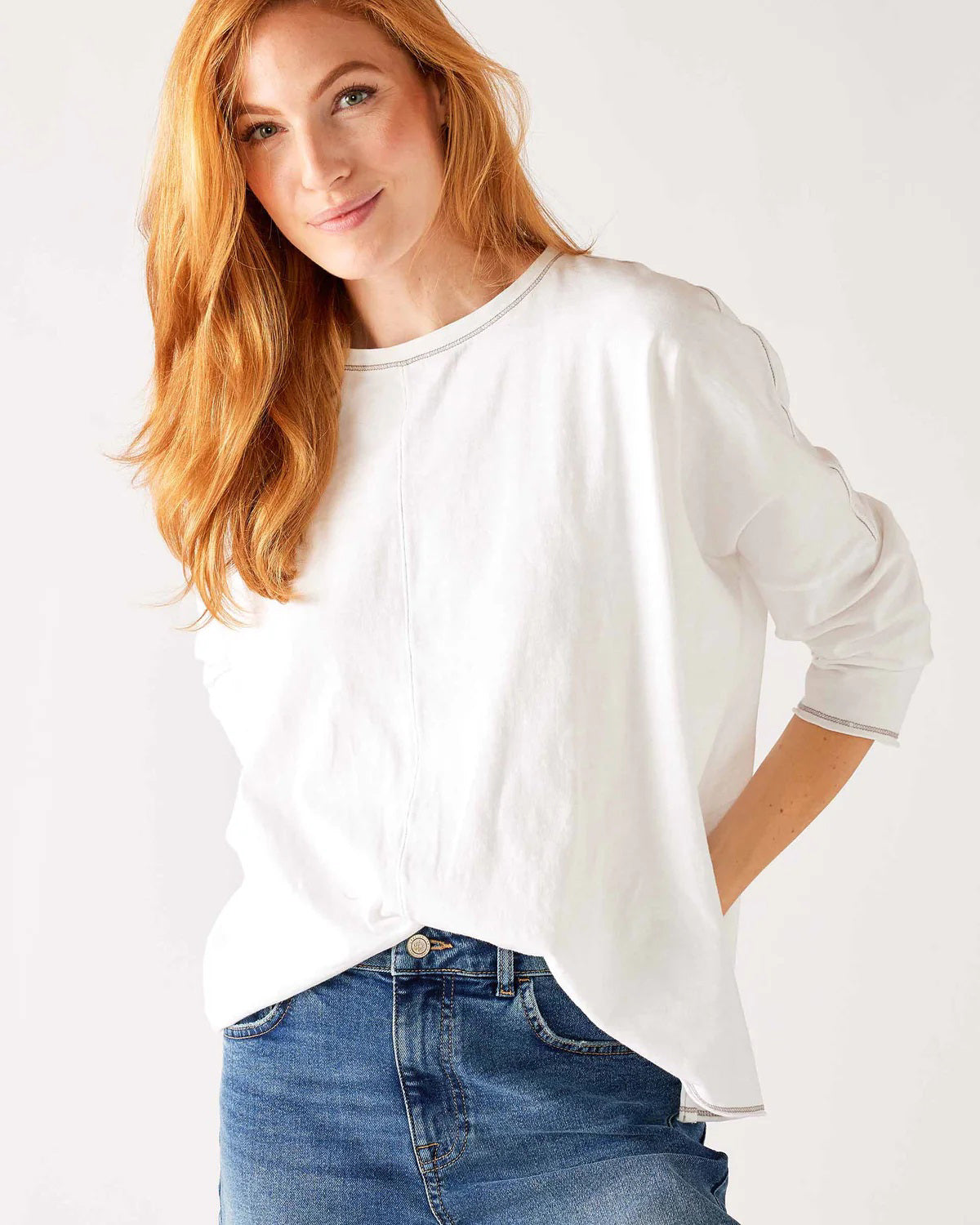 A woman with wavy brown hair models the MERSEA - CATALINA SLUB TEE—an oversized, long-sleeved white top with thin gray stripes—by MERSEA. She pairs it with blue jeans and stands against a plain, light background, hands behind her back.