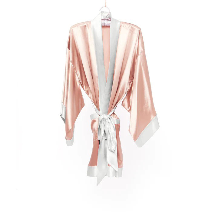 The BRANCHE GEISHA Robe Silk in pink satin, adorned with white trim, hangs gracefully on a matching silk hanger. This elegant robe by BRANCHE features long, wide sleeves and is accentuated with a white sash tied in a bow around the waist, complete with mitered corners for an exquisite finish.