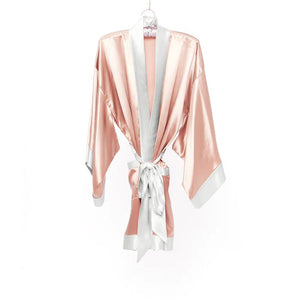 The BRANCHE GEISHA Robe Silk in pink satin, adorned with white trim, hangs gracefully on a matching silk hanger. This elegant robe by BRANCHE features long, wide sleeves and is accentuated with a white sash tied in a bow around the waist, complete with mitered corners for an exquisite finish.