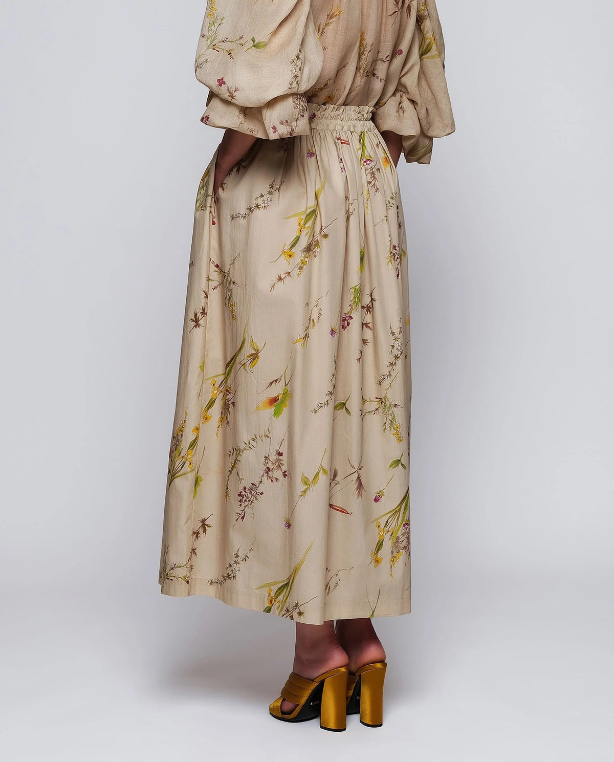 A woman stands against a plain background wearing the MIRTO 1956 Beige Floral Print Midi Skirt, paired with puffed three-quarter sleeves, a wide tan belt, and brown open-toe heels. Her hair is styled in a high bun with a decorative accessory.