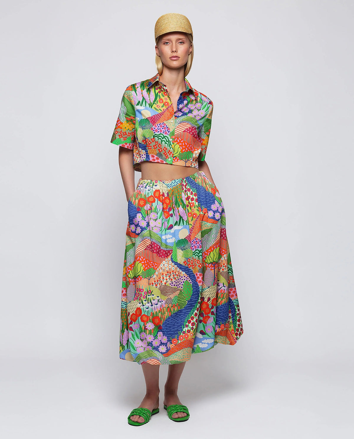 A woman stands against a plain background wearing green sandals and the MIRTO 1956 MULTICOLOR PRINT BALLOON SKIRT, featuring vibrant floral and peacock patterns, paired with a matching colorful crop top and straw hat.