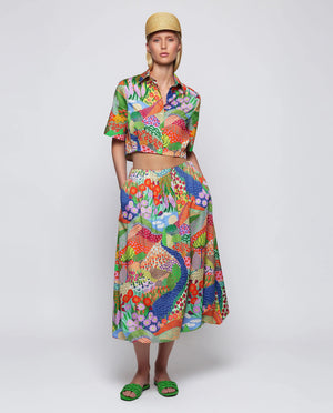 A woman stands against a plain background wearing green sandals and the MIRTO 1956 MULTICOLOR PRINT BALLOON SKIRT, featuring vibrant floral and peacock patterns, paired with a matching colorful crop top and straw hat.