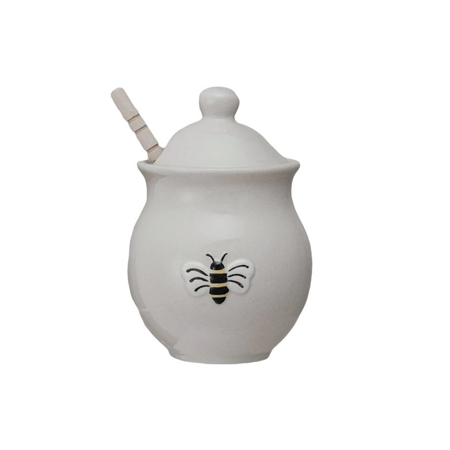 The HONEY JAR WITH EMBOSSED BEE AND WOOD DIPPER by CREATIVE COOP sits on a marble surface, partially covered by its lid. Honey oozes from the stoneware jar and pools beside it, with the wood dipper resting on top of the spill. The visually appealing setup conveys a sweet, natural theme.