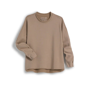 A woman with long brown hair stands indoors, her back to the camera, wearing the FRANK & EILEEN Anne Long Sleeve Capelet in beige and white pants near a wooden staircase and dining area with chairs.