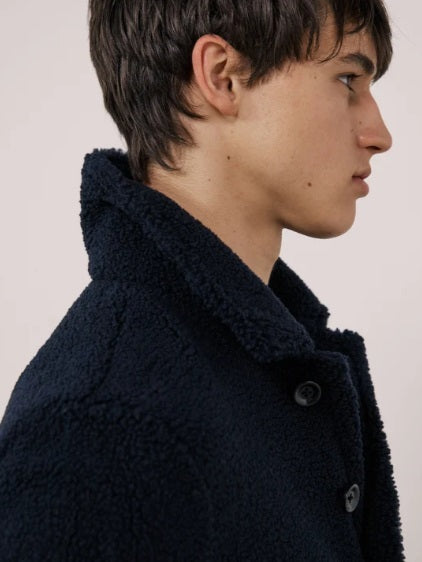 A young person with short dark hair wears the HARTFORD - Sherpa Utility Jacket in Navy by HARTFORD over a black shirt, looking directly at the camera against a plain light background. This warm sherpa layer brings both comfort and style to casual menswear.