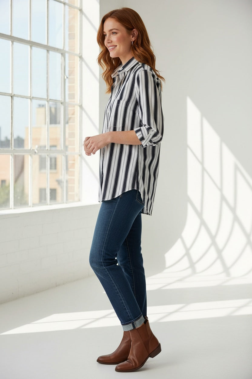 A woman stands outdoors in the sunlight, wearing the FRANK &amp; EILEEN Waverly NYC Button Up Shirt in Italian Brushed Superluxe Cotton with black and tan stripes. The shirt features rolled-up sleeves, and she is buttoning or unbuttoning its perfectly tailored fit.