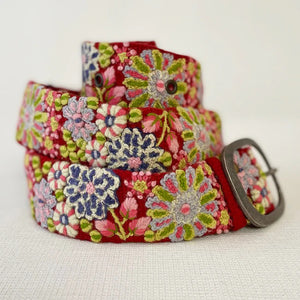 The JENNY KRAUSS - TEABERRY BELT by JENNY KRAUSS is a handcrafted, adjustable red belt with a silver buckle, featuring colorful embroidered floral patterns in pink, purple, green, blue, and white. Made in Peru.