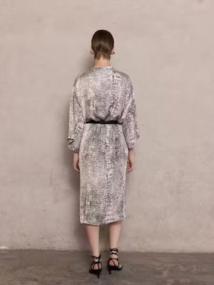 A woman stands against a beige wall, wearing the NUDE - ANIMALIER DRESS by NUDE. The dress features a loose, kaftan-inspired silhouette with black-and-white animal print, styled with wide-leg pants. Her hair is straight and pulled back, with a neutral expression.