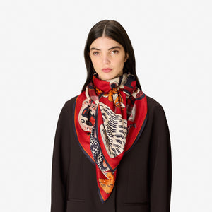 A woman with straight dark hair, dressed in a dark blazer, wears the luxurious INOUI EDITIONS - COLLECTIONNEUR SCARF WITH CATS IN MODAL SILK RED by INOUI. The red scarf—adorned with animal and wave designs and crafted from silk modal— is styled in a triangle around her neck as she stands against a plain white background.