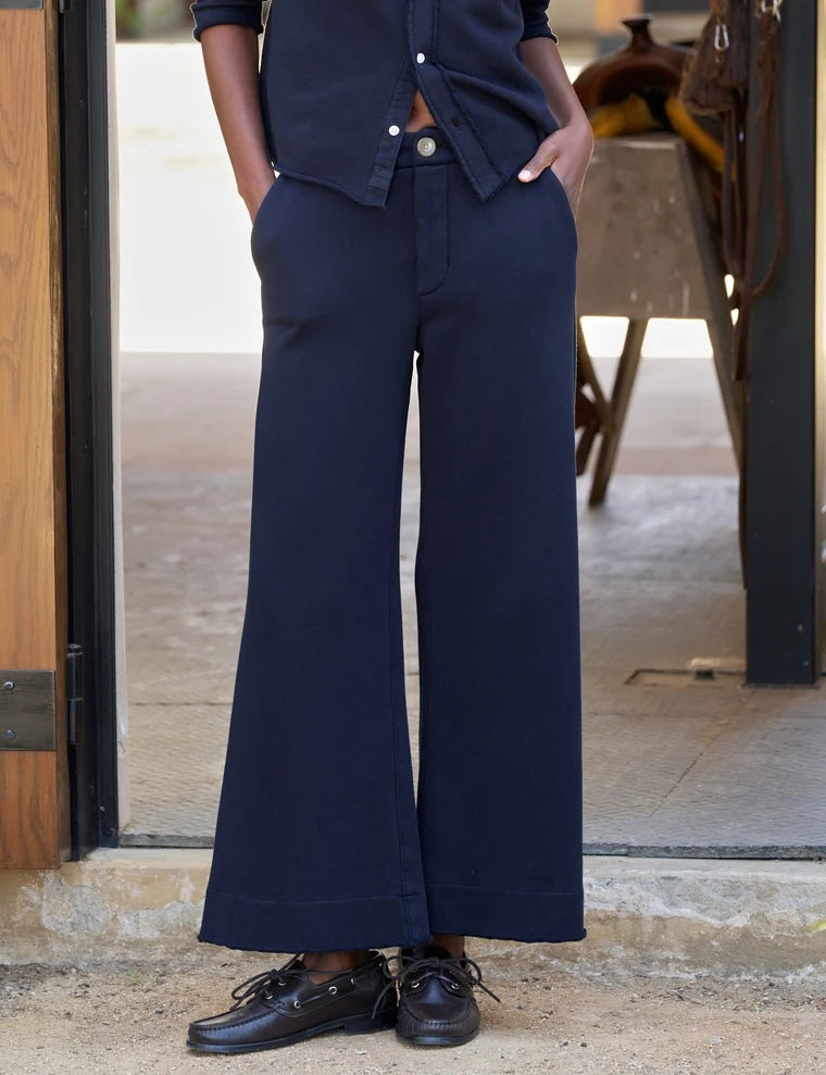A person stands outdoors facing away, wearing the FRANK & EILEEN Riley Trouser Sweatpant in Triple Fleece Classic Navy paired with black shoes, one hand resting by their side near a wooden wall and a bench—a relaxed look inspired by FRANK & EILEEN’s California Heritage Collection.