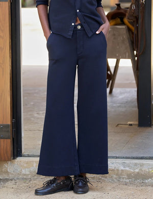 A person stands outdoors facing away, wearing the FRANK & EILEEN Riley Trouser Sweatpant in Triple Fleece Classic Navy paired with black shoes, one hand resting by their side near a wooden wall and a bench—a relaxed look inspired by FRANK & EILEEN’s California Heritage Collection.