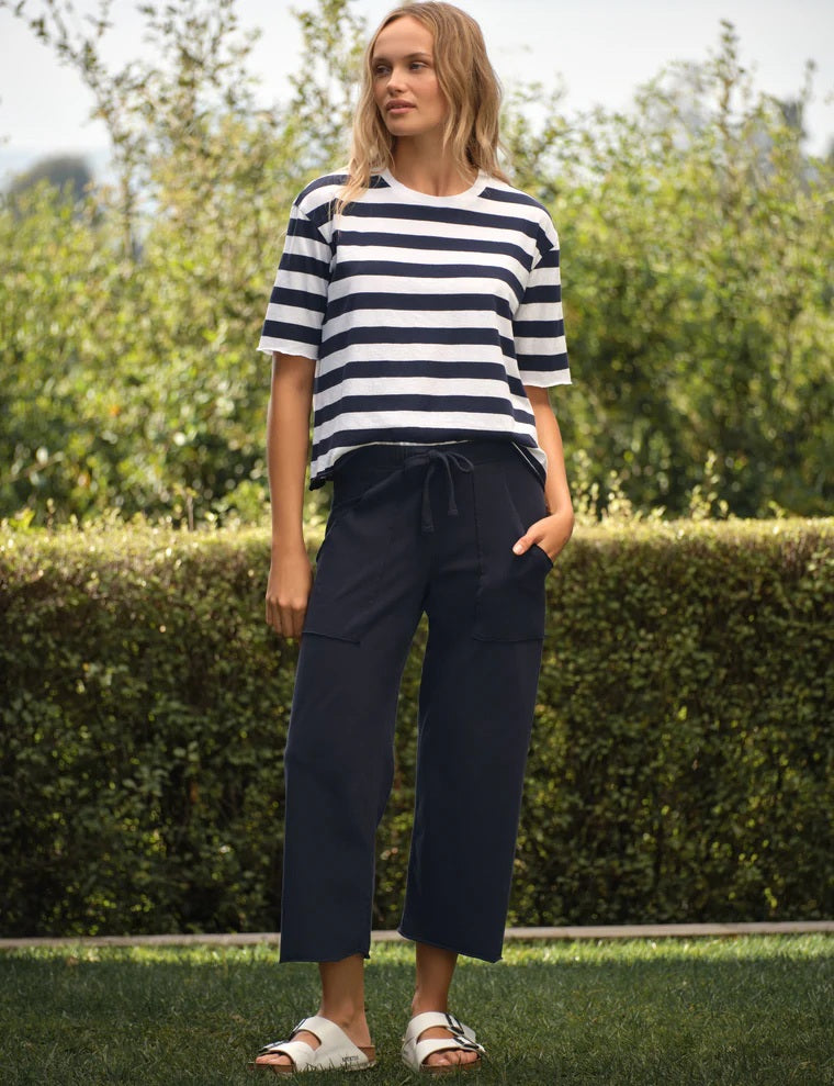A person wearing a striped shirt, white sandals, and the FRANK & EILEEN PENNY Patch Pocket Jersey Pant in British Royal Navy stands on grass with a hedge and greenery in the background.