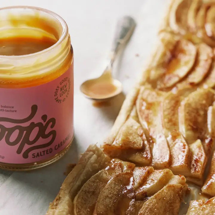 A jar of COOPS - SALTED CARAMEL SAUCE with pink labeling sits open, its thick, silky smooth sauce dripping down the side. The label reads "COOPS," boasting an all-natural, gluten-free treat with a perfectly sweet-salty balance and impossibly smooth texture.