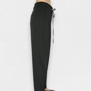 A person is wearing the MEIMEIJ - DRAWSTRING TROUSER PANT from the brand MEIMEIJ. These black, high-waisted trousers feature a loose fit and are made from a blend of polyester and elastane, complete with a tie belt. The image is taken from the waist down, showcasing the trousers in full against a plain white background, with the person's feet partially visible at the bottom.