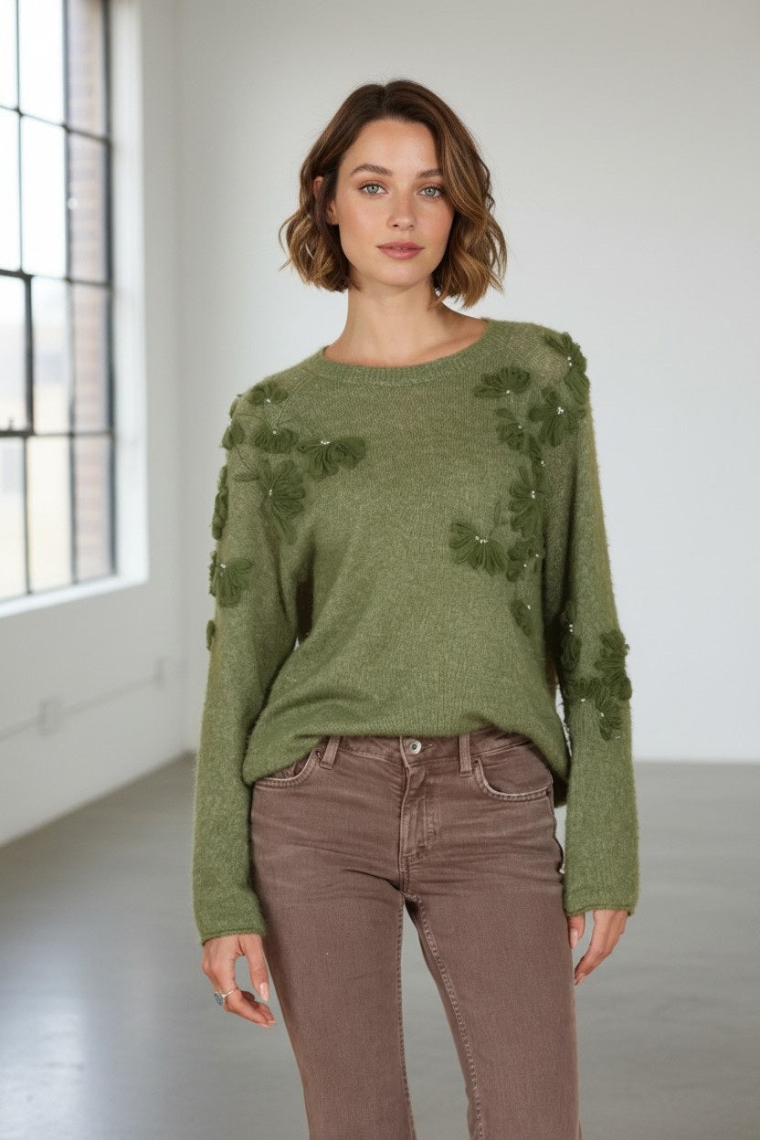 A woman with short, wavy hair stands indoors in front of large windows, wearing the JESSIE LIU Merino Wool and Cashmere Raglan Sleeve Pullover with Applique in Moss Green paired with brown pants. She faces the camera as natural light streams into the room.