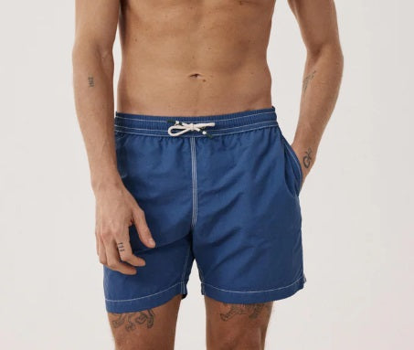 A shirtless person models the HARTFORD - NYLON SWIM TRUNKS WITH TRAVEL POUCH IN DENIM by HARTFORD against a plain light background, with tattoos visible on their arms and legs and one hand casually in a pocket.