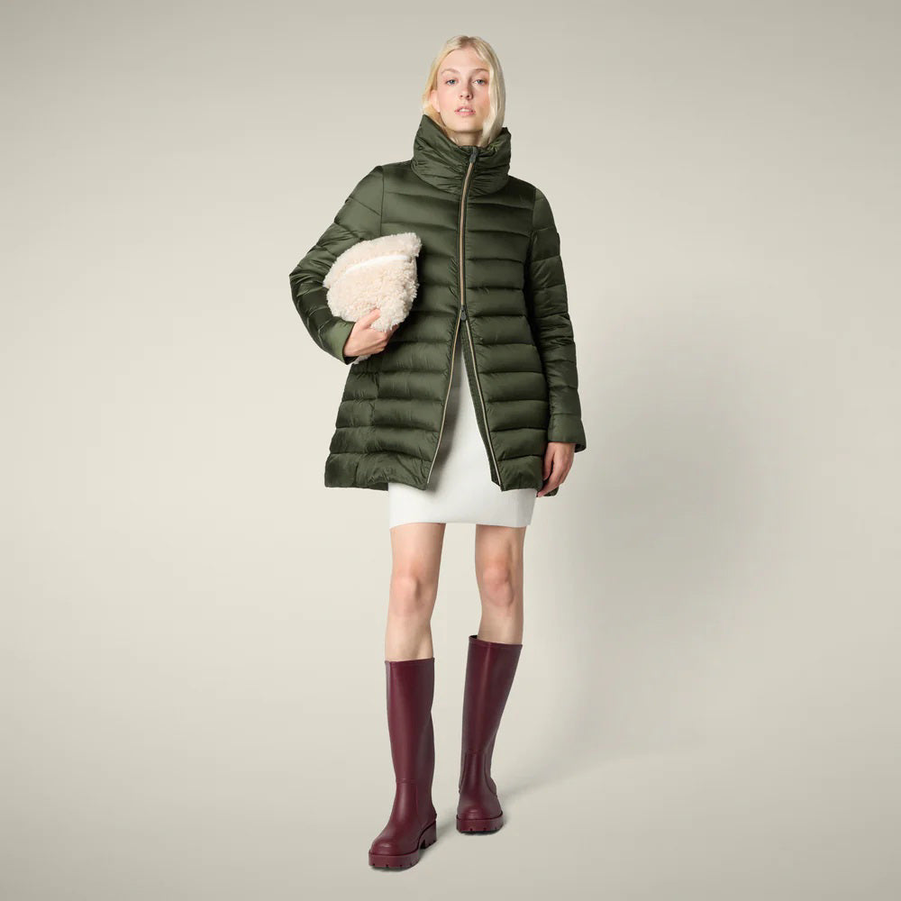 A woman stands against a plain light background in the SAVE THE DUCK - LYDIA PUFFER COAT, an olive green women&#39;s puffer jacket by SAVE THE DUCK, styled with a white dress, burgundy rain boots, and holding a fuzzy light pink hat.