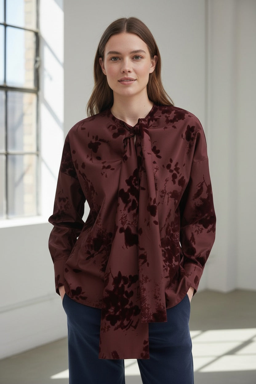 A woman stands indoors by a window, wearing the ROSSO35 taffeta and velvet top with a bow neckline in maroon from ROSSO 35 SILKY, paired with dark blue pants. She has long brown hair and a neutral expression.