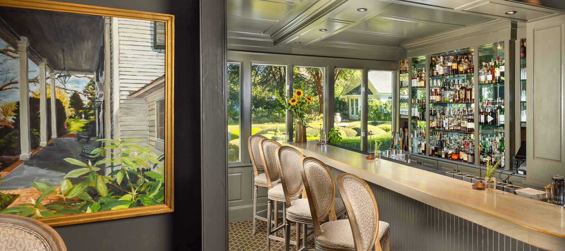 A stylish, well-lit home bar with a marble countertop, upholstered barstools, mirrored shelves stocked with liquor bottles, large windows overlooking a garden, and a framed painting of a porch on the adjacent wall.