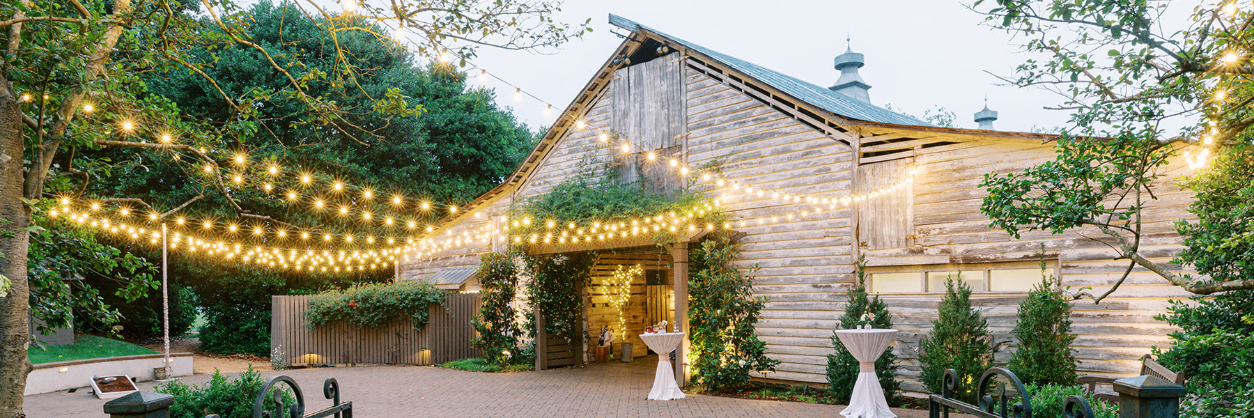 A rustic wooden barn decorated with strings of hanging lights, surrounded by trees and greenery. Cocktail tables with white tablecloths are set up outside on a paved area, creating a charming outdoor event space.