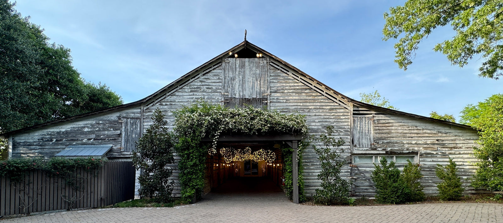 A large, rustic wooden barn with weathered gray boards stands under a blue sky. Ivy frames the entrance, which is lit inside by strings of warm, glowing lights. Green bushes and trees surround the building.