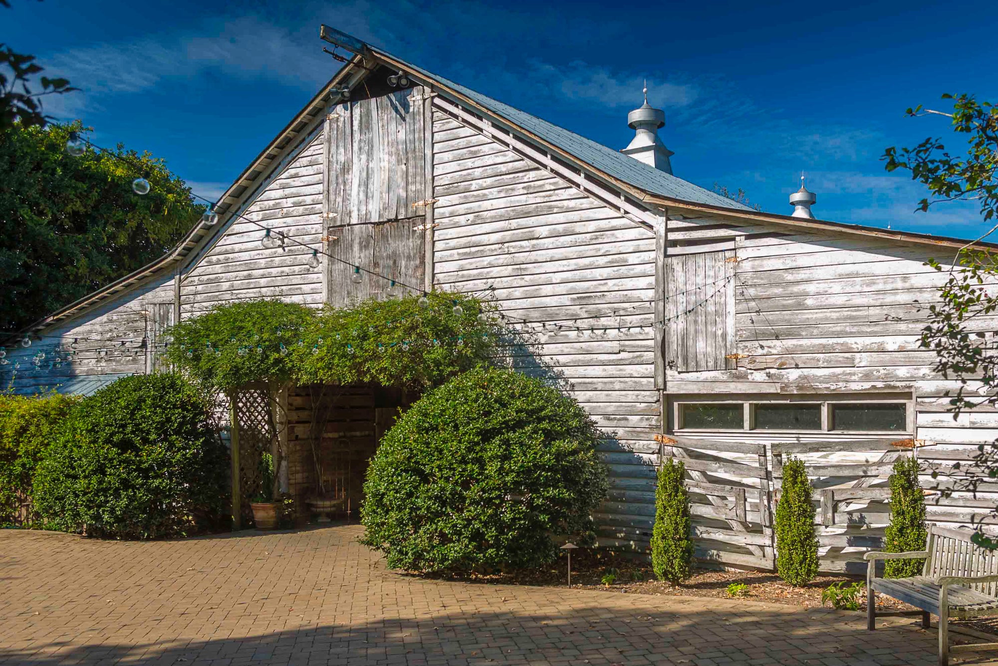 A weathered wooden barn with white peeling paint, surrounded by green bushes and trees, sits under a bright blue sky. The building has a sloped roof and a brick-paved area in front.