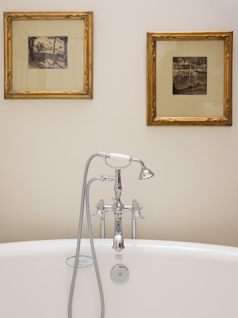 A white bathtub with a chrome faucet and handheld showerhead is positioned against a beige wall, below two framed black-and-white artworks.