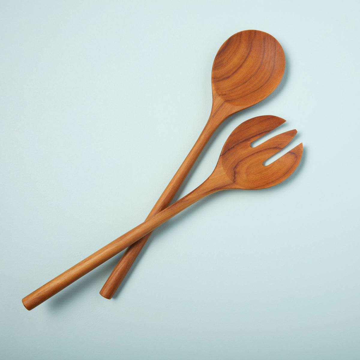 The BEHOME - TEAK SERVING SET by BE HOME, INC features two handcrafted teak servers—a spoon and a three-pronged fork—arranged crossed on a light blue background.