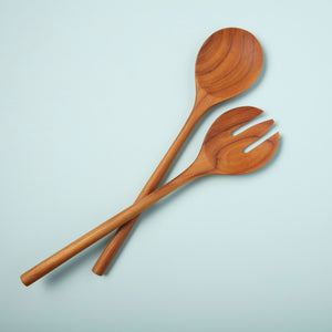 The BEHOME - TEAK SERVING SET by BE HOME, INC features two handcrafted teak servers—a spoon and a three-pronged fork—arranged crossed on a light blue background.
