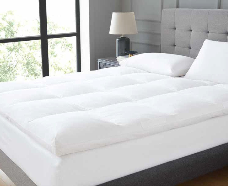 A neatly made bed with the DOWNLITE FEARRINGTON LIFESTYLE BEDDING COLLECTION - FEATHERBED QUEEN and accompanying pillows is positioned next to a bedside table featuring a modern black lamp. The room boasts a large window that allows natural light to brighten the space, emphasizing the grey upholstered headboard and light-colored walls.