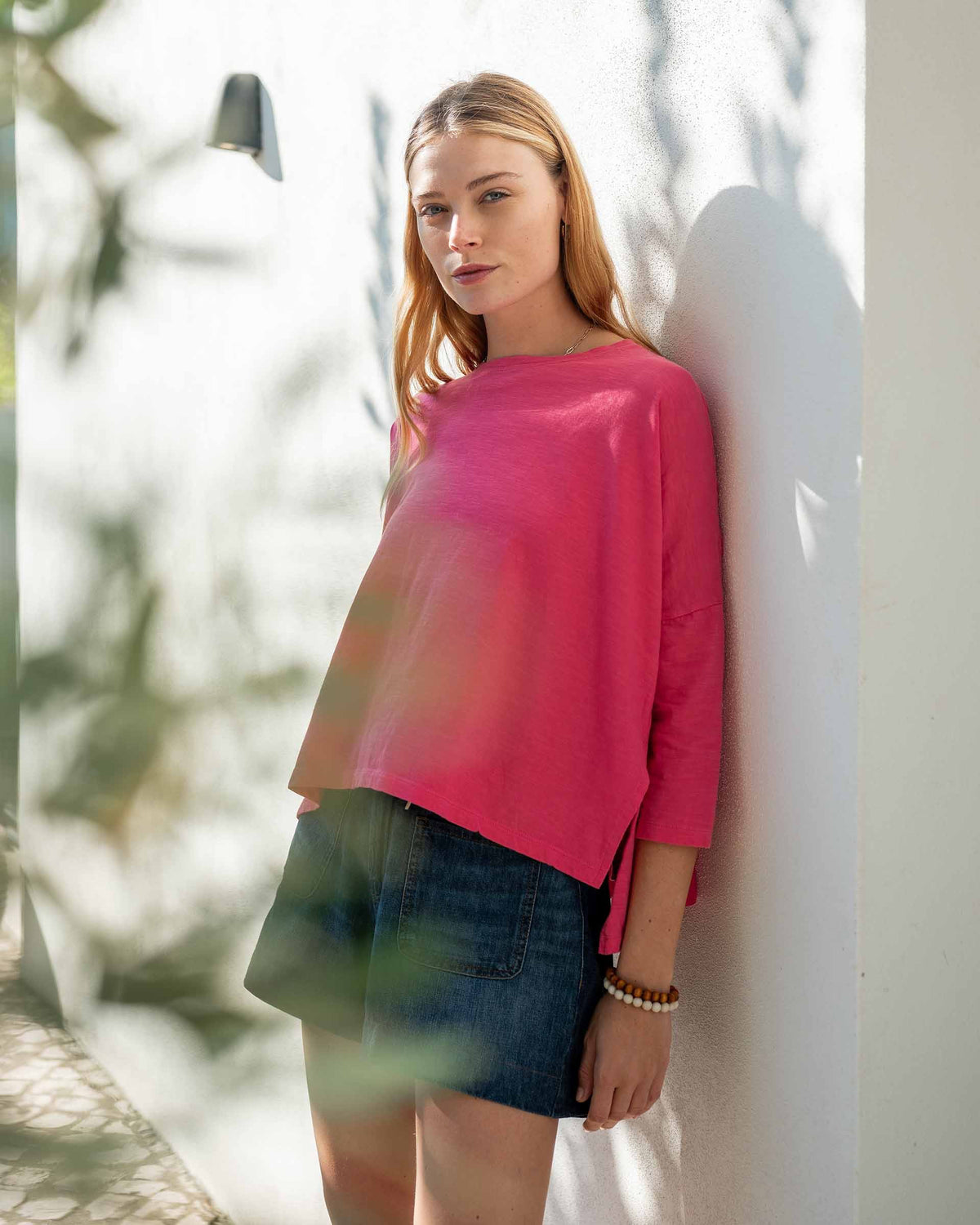 A woman with long, straight hair stands against a white wall, wearing the MERSEA Catalina Slub Tee in blue paired with white pants. The midweight textured cotton tee is from MERSEA. She looks at the camera with a neutral expression.