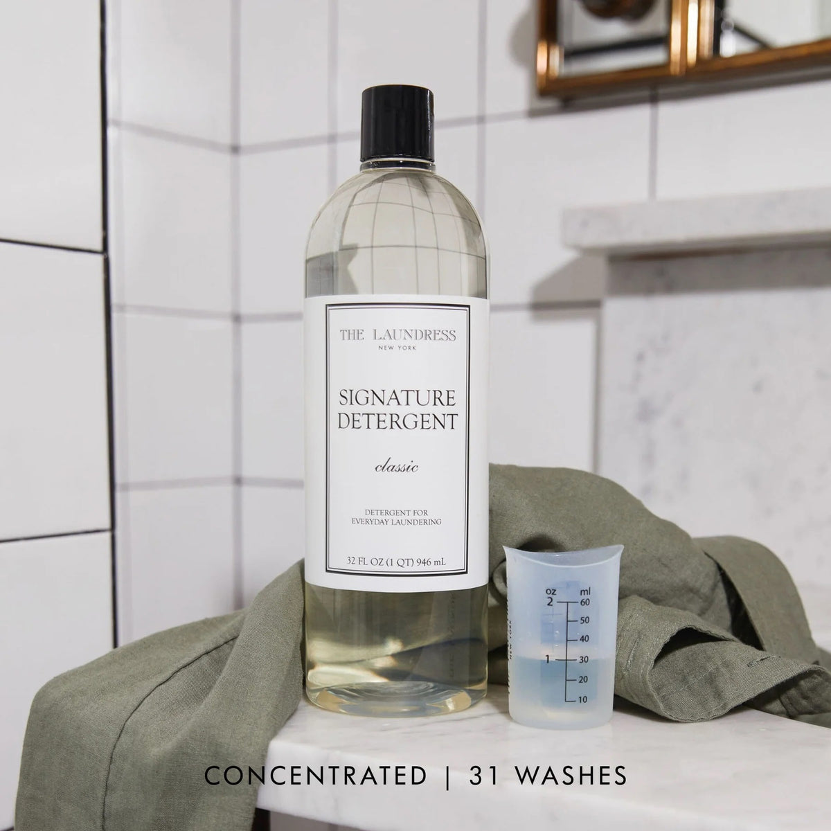 A 32 fl oz (946 ml) bottle of THE LAUNDRESS - SIGNATURE DETERGENT 32OZ featuring a classic scent and bio-based ingredients. The clear bottle comes with a black screw-top lid and a white label showcasing the product name and brand in black text. The background is a simple light gray.