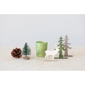 The CREATIVE COOP GARDEN KIT WITH HOLIDAY TREE features pine cones, green glittery candle holders, a bottle brush tree, and wooden cutouts of fir trees and a reindeer, all beautifully arranged on straw.