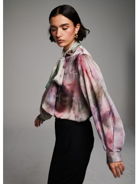 A woman with short dark hair wears the MILVA MI - BRUSHSTROKE BLOUSE by MILVA, a pastel long-sleeved statement bow top, paired with high-waisted black pants. She poses with eyes closed against a plain light background.