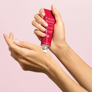 With perfectly manicured nails, a person applies MANUCURIST - COSMOS ORGANIC HAND CREAM, ROSE NOUVELLE from MANUCURIST INC to the back of their hand against a soft pink background, emphasizing the importance of caring for hands and nails.