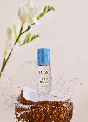 A LAFCO FINE FRAGRANCE, TROPIC WATERS 10ML box stands upright in a halved coconut, water splashing around it against a neutral background—an evocative display for this long-lasting scent inspired by the essence of coconut water.