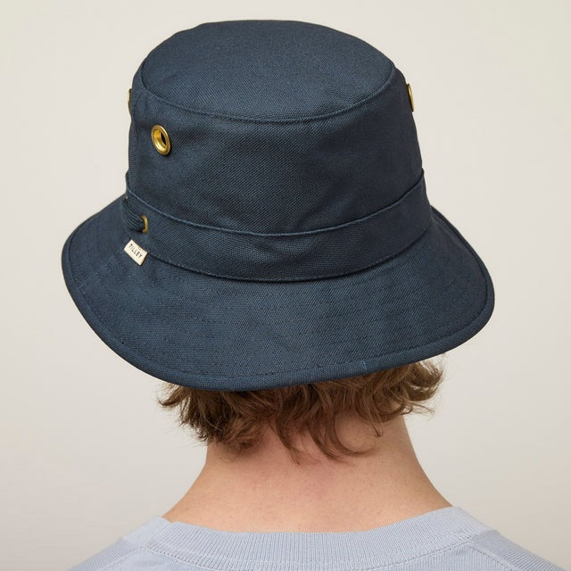A young person with fair skin and light brown hair wears the TILLEY - ICONIC T1 BUCKET HAT IN DARK NAVY and a light gray crew-neck shirt, looking directly at the camera against a plain, light background.