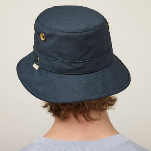 A young person with fair skin and light brown hair wears the TILLEY - ICONIC T1 BUCKET HAT IN DARK NAVY and a light gray crew-neck shirt, looking directly at the camera against a plain, light background.